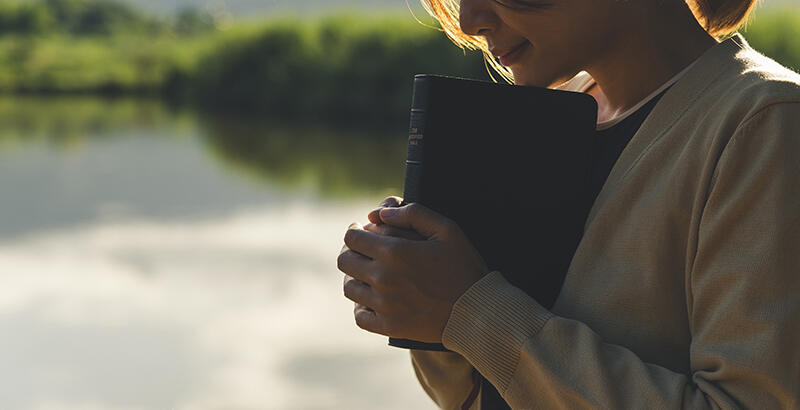A person holds a book against their chest beside still water at golden hour.