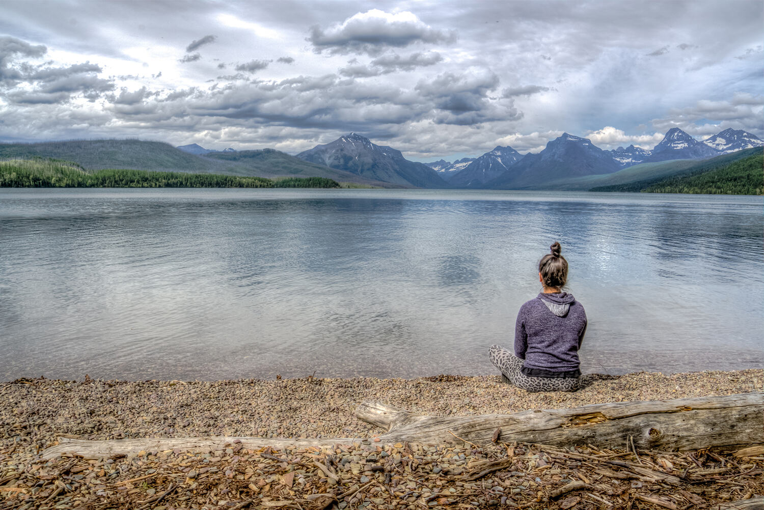 A woman sits alone on the shore of a still lake, looking out toward distant mountains.