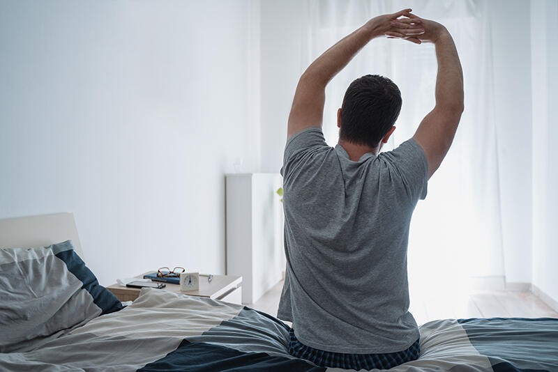 A man sits on the edge of his bed and stretches, morning light at the window.
