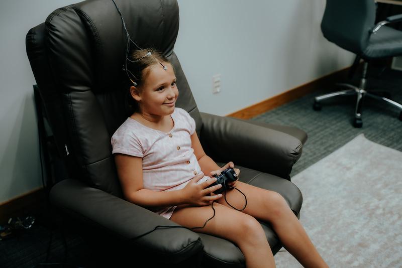 A young girl sits in the neurofeedback chair, sensors in her hair, holding a games controller.