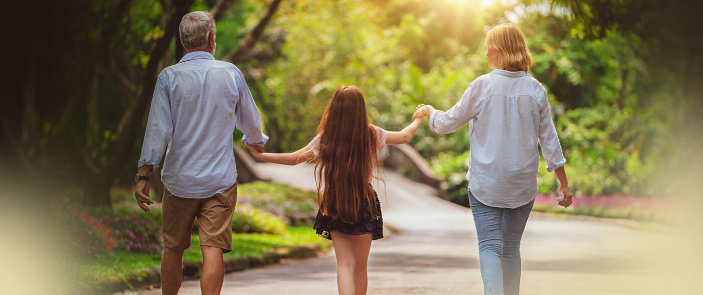 A family walks away down a sunlit park path, holding hands.