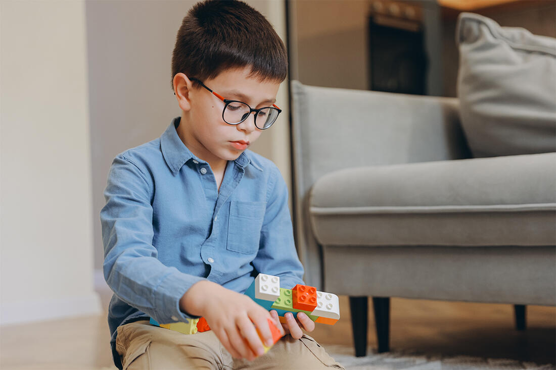 A young boy in glasses sits on the floor at home, absorbed in building with plastic bricks.