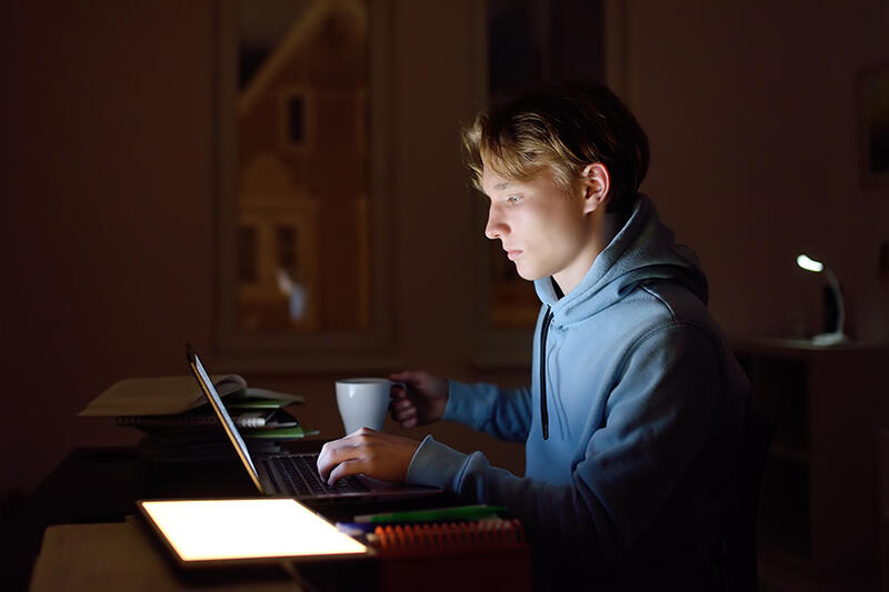 A teenage boy works at a laptop late at night, a mug in one hand.