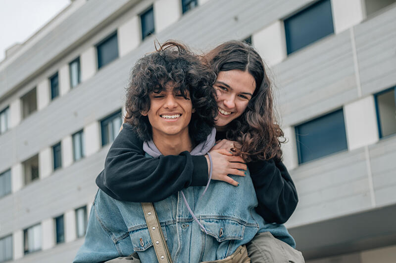 Two teenagers outdoors, one hugging the other from behind, both laughing.