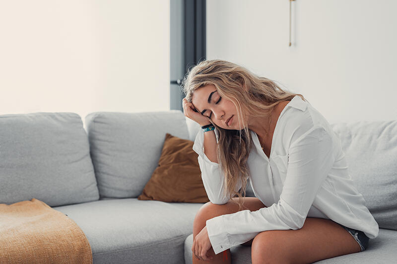 A woman sits alone on a sofa, her head resting in her hand.