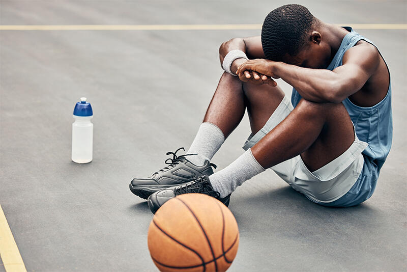 A young basketball player sits on the court with his head down, the ball beside him.
